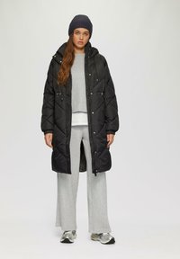 Langer schwarzer Daunenmantel mit Kapuze, mit einem gesteppten Design, Druckknöpfen und Kordelzug. Getragen über einem grauen Pullover und gemusterten Hosen.