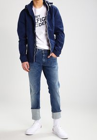 Giacca blu navy con cappuccio e cordini bianchi, t-shirt bianca con grafica, jeans azzurri con risvolti e scarpe da ginnastica bianche con dettagli rossi.