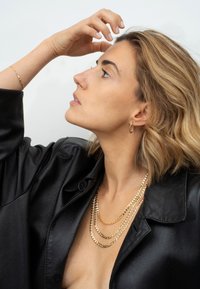 Goldfarbene geschichtete Halsketten und Creolen, getragen mit einem schwarzen Satinhemd, das die glatte Textur und den kontrastierenden Glanz zur Schau stellt.