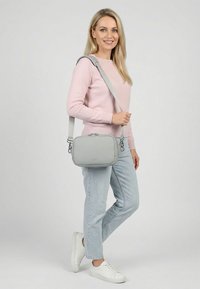 Lächelnde Frau mit rosa Pullover und hellen Jeans trägt eine hellgraue Umhängetasche und steht mit einem leicht angehobenen Fuß.