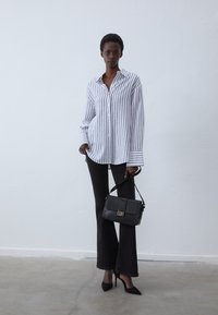 Striped button-up shirt in white and blue, paired with black flared pants. Model holds a textured black handbag with a gold clasp.