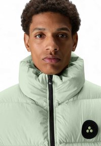 Mintgrüne gesteppte Jacke mit hohem Kragen, ausgestattet mit einem schwarzen Reißverschluss und einem runden Logo-Patch auf der Brust. Glatte Textur.