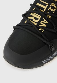 Botte de sport noire avec un dessus texturé, un texte doré en relief, des lacets noirs épais et une accentuation rayée blanche contrastante sur la semelle.
