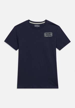 Temno modra majica s kratkimi rokavi z majhno belo značko z logotipom "GUESS" na zgornjem levem delu prsnega koša in okroglim izrezom