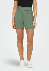 Calções verdes, leves, com uma cintura franzida, combinados com sapatilhas brancas. O tecido tem uma textura suave e um corte descontraído.