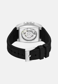 Tylnia część zegarka ze stali nierdzewnej w kolorze srebrnym, z widokiem na mechanizm; czarny gumowy pasek z regulowanym zapięciem i tłoczonym wzorem.