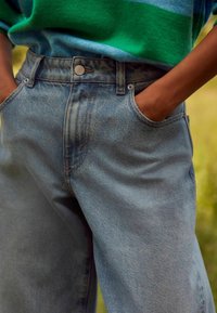 Blugi din denim albastru deschis cu talie înaltă, prezentând buzunare laterale proeminente și un închizător vizibil cu buton. Textura ușor decolorată accentuează adâncimea.