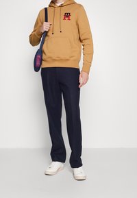 Uomo che indossa una felpa beige con un logo rosso e nero, pantaloni blu, sneakers bianche e una borsa a tracolla blu con un motivo rosso.