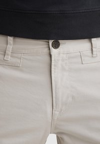 Nahaufnahme der Taille einer beigen Hose mit Gürtelschlaufen, Metallknopf und einem Teil eines schwarzen, gerippten Hemdes, das eingesteckt ist.