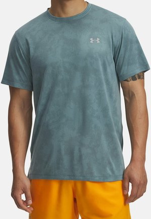 Mężczyzna w jasnoniebieskiej koszulce Under Armour w kamuflaż i jaskrawych pomarańczowych szortach sportowych, ramiona swobodnie opuszczone wzdłuż ciała.