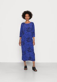 Vestido longo azul com padrão de respingos pretos, gola redonda, mangas três-quartos e fendas laterais. Combinado com mules pretas de salto bloco.