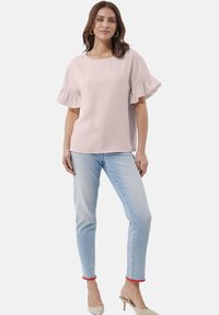 Hellrosa Bluse mit Rüschensäumen, rundem Ausschnitt, kombiniert mit hellblauen Jeans. Die Model trägt beige Absatzschuhe mit roten Pom-Pom-Akzenten.