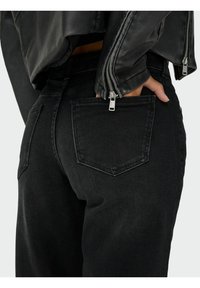 Črne denim hlače z ozkim krojem, s hrbtnimi žepi in šivanim detajlom. Nosi se z črno usnjeno jakno, ki prikazuje teksturirano tkanino.