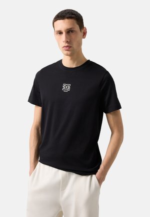 Młody mężczyzna w czarnej koszulce z krótkim rękawem z małym białym logo na piersi i białych spodniach, ręce w kieszeniach, na jednolitym tle.