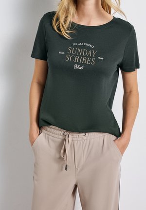 Persona con camiseta de manga corta verde oscuro con el texto "Sunday Scribes Club" y pantalones beige con cordón, manos en los bolsillos, de pie frente a un fondo liso.