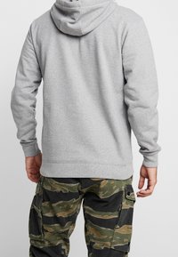 Haut à capuche gris avec cordon de serrage, fabriqué en mélangé de coton doux. Présente des poignets et une taille côtelés, porté avec un pantalon cargo camouflage.