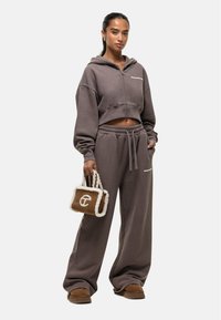 Geknipte bruine hoodie met rits, losse bijpassende trainingsbroek en een kleine bruine tas met beige nepbontafwerking en logodetail. Zacht materiaal.