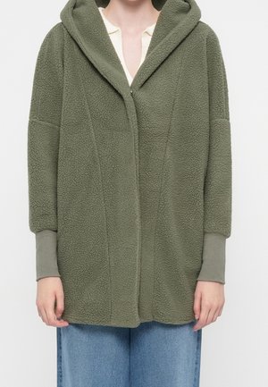Personne portant une veste polaire vert olive zippée à capuche avec des poignets côtelés et un jean bleu, debout devant un fond blanc.