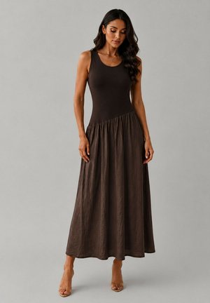 Femme aux longs cheveux foncés portant une robe maxi marron foncé sans manches avec un haut ajusté et une jupe froncée, debout devant un fond gris uni.