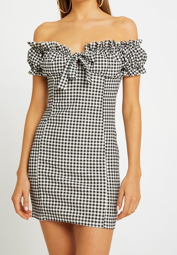 Femme portant une mini-robe noire et blanche à carreaux vichy, épaules dénudées, manches bouffantes et détail noué à l'avant.