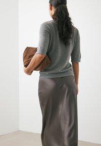 Maglione grigio lavorato a maglia con maniche corte, abbinato a una pochette in tessuto marrone, indossato con una gonna lunga, liscia e di satin scuro.