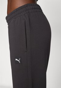 Schwarze Sporthose aus weichem Stoff, mit einem elastischen Bund und einer Seitentasche. Ein weißes Puma-Logopatch ist an dem unteren Bein sichtbar.