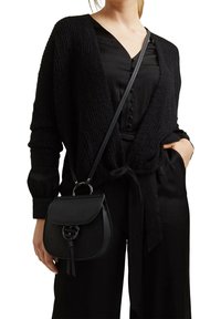 Bolso bandolera negro con forma redondeada, acabado texturizado, herrajes de cierre prominentes y un adorno de borlas, combinado con un atuendo negro.