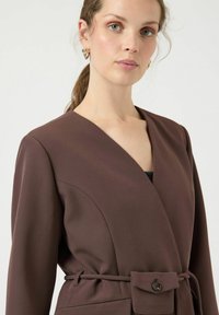 Bruine wikkeljas met een V-hals, lange mouwen, een tailleband en een voorzak met knoopsluiting. Gladde stof met een gestructureerd ontwerp.