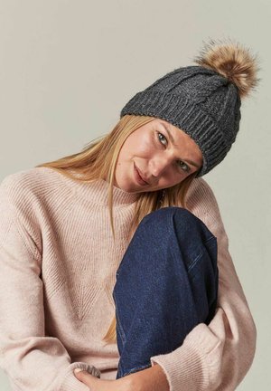 Bonnet en tricot gris avec un motif texturé et un pompon en fausse fourrure sur le dessus, porté avec un pull rose clair et un jean bleu foncé.