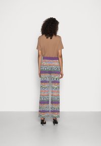 Hnědé tričko, vzorované široké kalhoty s fialovými, oranžovými a modrými akcenty. Zadní pohled ukazuje elastický pas a volný design.