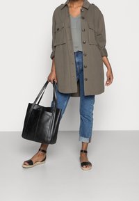 Camisa chaqueta de gran tamaño en verde oliva con cierre de botones y dos bolsillos delanteros, combinada con jeans azules con puños, bolsa de mano de cuero negro y sandalias negras.