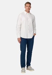 Camicia bianca in cotone a maniche lunghe con colletto a punta, bottoni frontali, abbinata a jeans in denim blu scuro e sneakers beige.