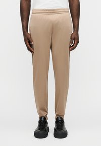 Μπεζ αθλητικό παντελόνι με ελαστική μέση, στενόμακρα πόδια και λεία υφή. Φορεμένο με μαύρα αθλητικά παπούτσια με στρογγυλές μύτες.