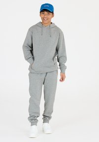 Grijze hoodie met trekkoord, gecombineerd met grijze joggingbroek. Bevat elastische manchetten en een elastische tailleband. Draag het met witte sneakers en een blauwe pet.