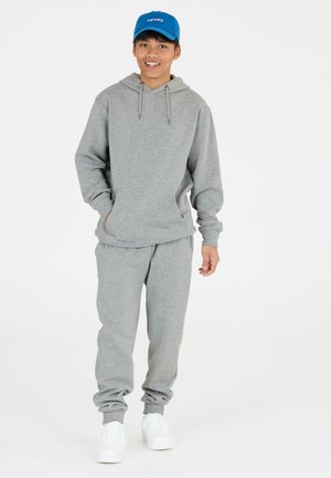 Grå hoodie med dragsko, kombinerad med grå sweatpants. Har elastiska muddar och midja. Bärs med vita sneakers och en blå keps.