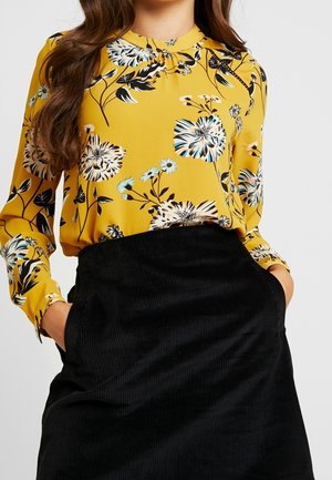 Femme portant un chemisier floral jaune à manches longues rentré dans une jupe en velours côtelé noire taille haute, les mains dans les poches.