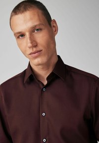 Burgunderfarbendes Hemd mit Knopfleiste, subtiler Struktur, spitzem Kragen und kontrastierenden blauen Knopfakzenten. Taillierte Passform, keine sichtbaren Muster.