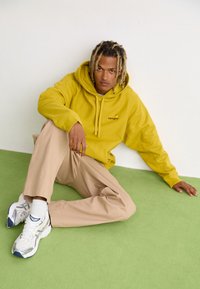 Haut à capuche en polaire jaune avec cordons de serrage et petit logo, associé à un pantalon large beige. Baskets blanches avec accents argentés. Sol vert.