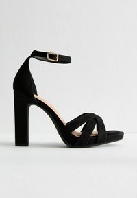 Svart mocka högklackad sandal med korsbanddesign, ankelrem, fyrkantig tå och en kraftig klack. Guldspänne som accent på remmen.