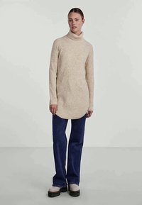 Suéter bege de gola alta e canelado com barra alongada, combinado com jeans azuis de perna larga e botas de sola grossa.