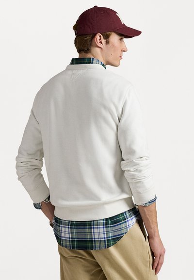 Ung mann iført en kastanjebrun caps, hvit sweatshirt over en grønn-blå rutete skjorte, og beige bukser, som står med hendene i lommene, vendt bort.