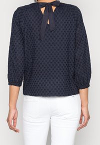 Blusa blu navy a texture con maniche a tre quarti, dotata di una chiusura a fiocco sul retro e polsini arricciati, indossata con pantaloni bianchi.
