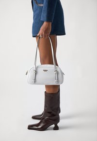 Bolso de cuero blanco con dos asas cortas, herrajes plateados, diseño redondeado y cierre con cremallera. Posicionado sobre un fondo neutro.