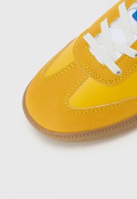 Żółty tenisówka z białymi sznurowadłami, zamszowymi i skórzanymi panelami oraz brązową gumową podeszwą na białym tle.