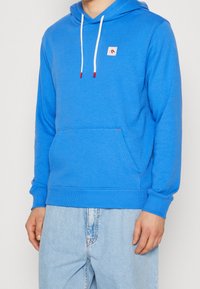 Sudadera azul hecha de una tela suave, con un bolsillo frontal, cordones blancos y un pequeño parche de logo en el pecho.