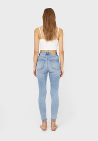 Mujer con cabello largo y castaño, vistiendo una camiseta blanca corta, jeans ajustados de color azul claro y sandalias de tiras color canela, de pie de espaldas.