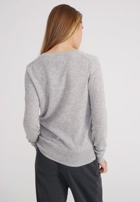 Grauer Strickpullover mit Rundhalsausschnitt und langen Ärmeln, der eine strukturierte Oberfläche und einen gerippten Saum aufweist. Die Rückseite ist etwas länger als die Vorderseite.