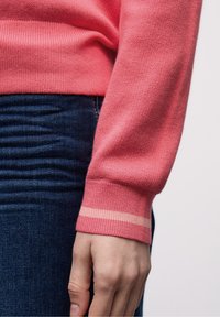 Gros plan d'une main reposant à côté d'un jean bleu foncé, portant un pull à manches longues rose avec une bande rose plus claire au niveau du poignet.