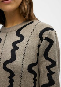 Bežinis megztinis su juodu banguotu raštu ir taškeliais. Jo tekstūra atrodo minkšta, su apvaliu kaklu ir vidutiniu pritaikymu.