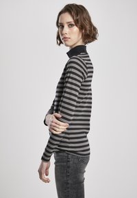 Langærmet grå og sort stribet top med høj hals, lavet af et blødt materiale. Båret med mørkegrå jeans. Modellen står sidelæns.